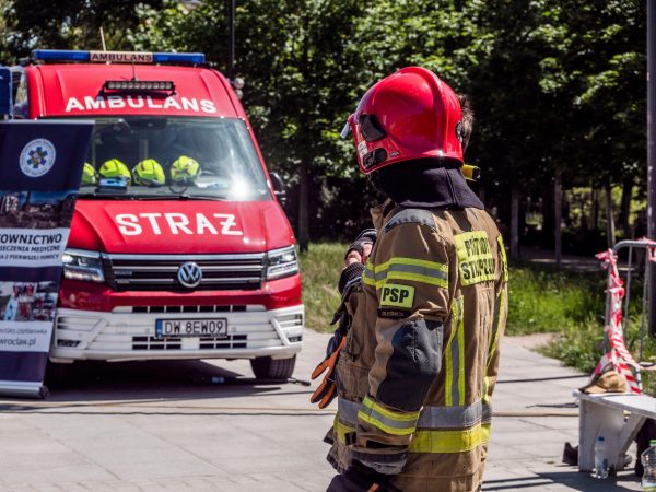 Firefighter in action near red ambulance labeled 'STRAŻ' outdoors in a daytime emergency scene.